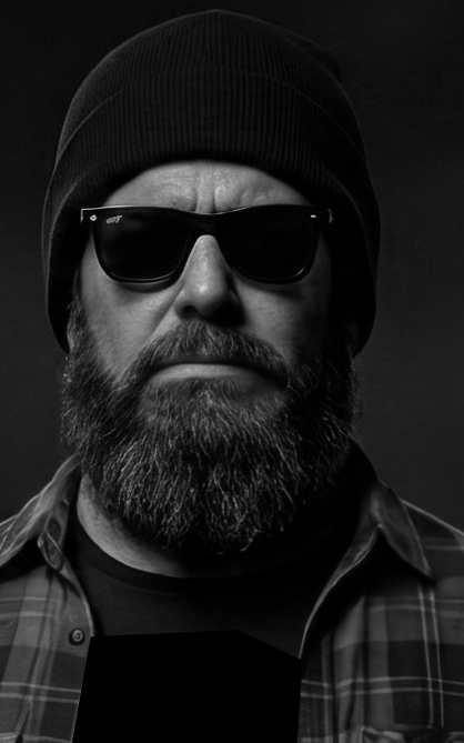 imagem em preto e branco de um homem com barba e óculos escuros, usando um gorro e camisa xadrez, fundo escuro